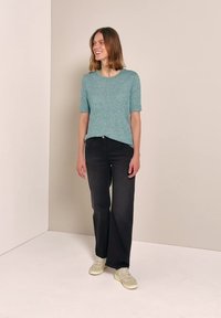 Kvinde, der står i et hjørne iført en teal kortærmet sweater, sorte bukser med vide ben og hvide sneakers, smilende og kigger til siden.