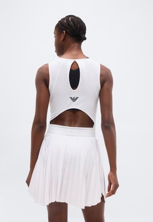 Donna con capelli intrecciati che indossa un top bianco senza maniche con incavi e una gonna bianca plissettata su uno sfondo semplice.