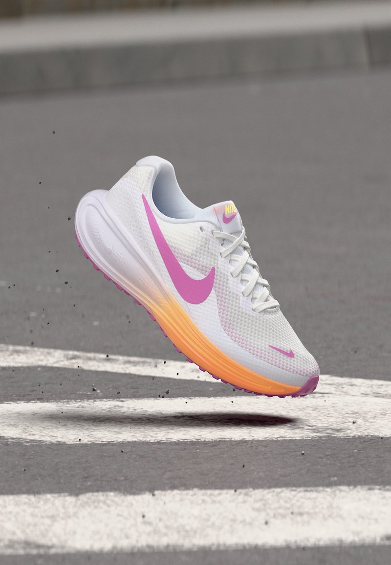 Λευκό αθλητικό παπούτσι Nike με ροζ σήμα και πορτοκαλί σόλα αιωρείται πάνω από γκρίζο πεζοδρόμιο με λευκές γραμμές.