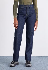 Jeans bleumarin cu talie înaltă și siluetă dreaptă. Cusături contrastante și închidere cu buton în față. Purtați cu adidași gri.