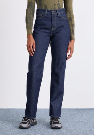 Donkerblauwe hoge taille jeans met een rechte pijp. Contrasterende stiksels en sluiting met een knop aan de voorkant. Draagt met grijze sneakers.