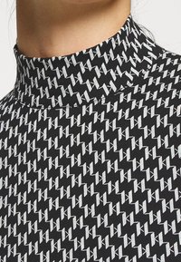 Tessuto nero con un motivo geometrico bianco ripetuto e un design a colletto alto. La texture appare liscia e leggermente elasticizzata.