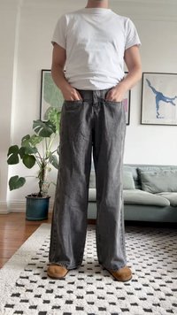 Jeans de pierna ancha de color gris oscuro con un corte recto, que cuentan con bolsillos frontales y una cintura alta. Combinados con una camiseta blanca y calzado marrón.