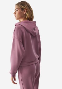 Sudadera con capucha en suave tela malva. Presenta un ajuste relajado, hombros caídos y puños acanalados. Combinada con pantalones a juego.