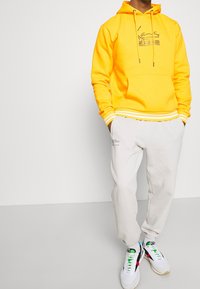Sudadera amarilla con capucha, bolsillo frontal, puños y dobladillo acanalados. Combinada con pantalones de chándal gris claro y zapatillas blancas con acentos coloridos.