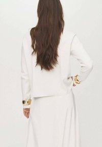 Maglione bianco corto con maniche lunghe, dettagli in oro sui polsini e tessuto textured. I capelli cadono in onde lungo la schiena, incorniciando il capo.