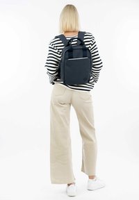 Námořnický materiál batoh s dvojitými ramenními popruhy, minimalistický design, přední kapsa a madlo na vrchu. Model má na sobě pruhovanou košili a béžové široké kalhoty.