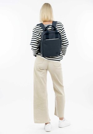 Zaino in tessuto blu marine con doppie spalline, design minimalista, tasca frontale e manico superiore. Il modello indossa una camicia a righe e pantaloni larghi beige.