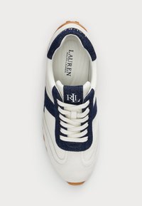 Bílé tenisky s námořnickými modrými akcenty, které mají mix texturované tkaniny a hladkého materiálu, kulatou špičku a bílé tkaničky. Logo viditelné na jazyku.