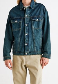 Spijkerjasje in een vervaagde donkerblauwe kleur, met twee borstzakken, zilveren knopen en een klassieke kraag. Draagt over een licht overhemd en kaki broek.