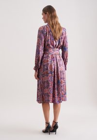Lila- und rosa-gemustertes Paisley-Kleid mit langen Ärmeln, gerafftem Rücken und einem passenden Gürtel. Tragen mit schwarzen, hochhackigen Sandalen.