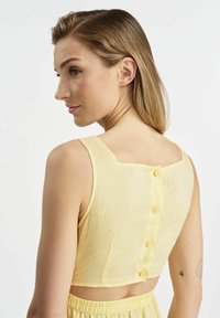 Żółty krótki top wykonany z teksturowanej tkaniny, z kwadratowym tyłem i sześcioma pasującymi guzikami. Bez rękawów, o dopasowanej sylwetce.