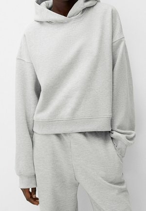 Person wearing a light gray hooded sweatshirt with long sleeves and matching sweatpants, hand in pocket.