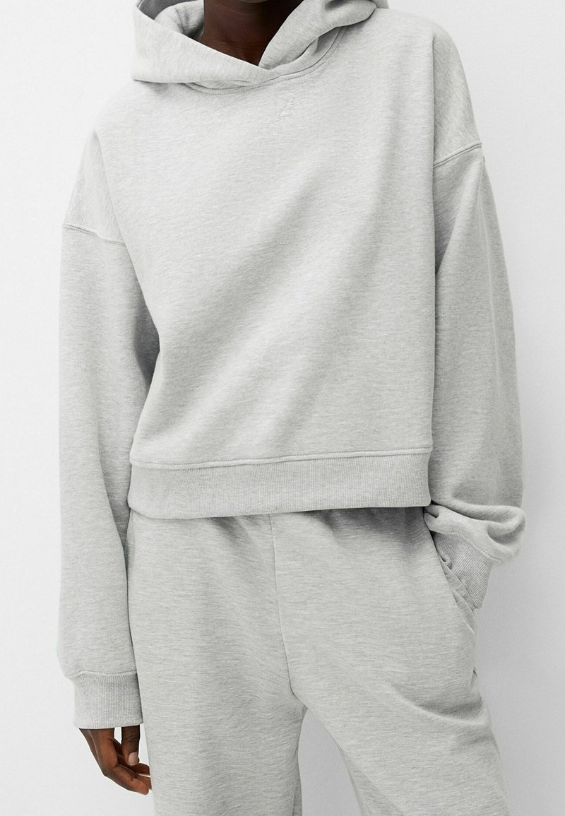 Personne portant un sweat à capuche gris clair à manches longues et un pantalon de survêtement assorti, avec la main dans la poche.