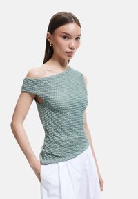 Top off-the-shoulder en tejido texturizado de color verde claro con detalle fruncido. Ajuste slim, mangas cortas, combinado con pantalones blancos holgados.