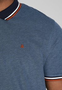 Blue polo shirt with a textured weave, featuring a navy collar and orange trim. Small orange logo is centered on the chest.