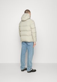 Beige puffjacka med huva, tjock vadderad design med breda horisontella paneler, bärs över ljusblå denimbukser och svarta skor.
