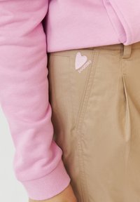 Lichtroze sweatshirt met geribbelde manchetten, gecombineerd met tan broek die een klein geborduurd hartje en de tekst "DOGETHER" nabij de zak heeft.