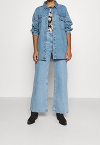 Persona che indossa un giubbotto di jeans azzurro chiaro e jeans a gamba larga, top floreale e stivaletti neri alla caviglia, in piedi davanti a uno sfondo bianco semplice.