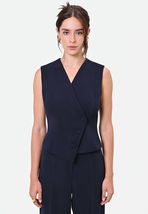 Donna con capelli scuri in un gilet blu navy senza maniche con tre bottoni e pantaloni abbinati, che indossa orecchini d'oro e un anello.