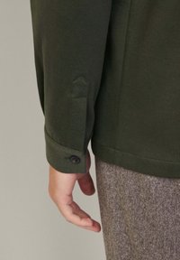 Camicia verde a maniche lunghe con polsini abbottonati, esposta con una mano appoggiata alla cucitura; tessuto texturizzato e design su misura.