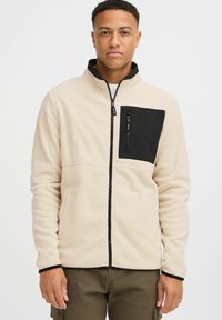 Chaqueta de forro polar beige con un bolsillo en el pecho con cremallera negra, cuello alto y acentos negros en los puños y la cremallera. Textura suave y diseño ajustado.