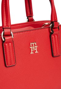 Κόκκινη δερμάτινη τσάντα με υφή, διπλές λαβές και ένα μικρό χρυσό μεταλλικό έμβλημα λογότυπου στο μπροστινό πάνελ.