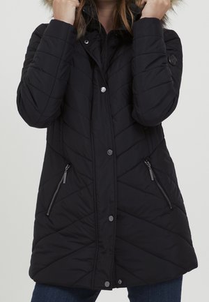 Femme portant un manteau d’hiver matelassé noir avec une capuche bordée de fourrure, tenant la capuche près de son visage.