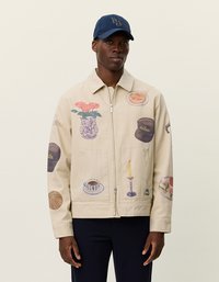 Μπεζ μπουφάν με διάφορες εκτυπωμένες εικονογραφήσεις, περιλαμβανομένων λουλουδιών, τροφών και ενός κεριού, με δύο μπροστινές τσέπες.