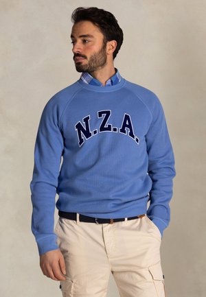 Man met baard, gekleed in een lichtblauwe N.Z.A. trui, een blauw geruit overhemd, beige cargobroek en een zwarte riem, staand met handen in de zakken.