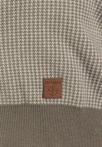 Gebreide trui met een houndstoothpatroon in beige en bruin, voorzien van een bruin leren label met "KEY LARGO" aan de onderkant.