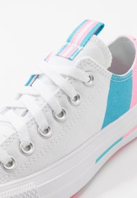 Zapatilla blanca de lona con rayas diagonales en azul y rosa, parte delantera con cordones, ojetes plateados y un tirador en el talón a rayas para un detalle adicional.