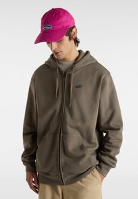 Brauner Reißverschluss-Hoodie mit Kordelzug und zwei Fronttaschen. Kombiniert mit einer leuchtend pinken Mütze mit blauem Logo. Glatte Textur.