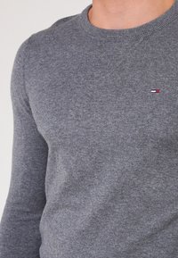 Šedý pletený svetr s kulatým výstřihem a žebrovanými manžetami, s malým logem vyšitým na hrudi v červené, bílé a modré barvě.