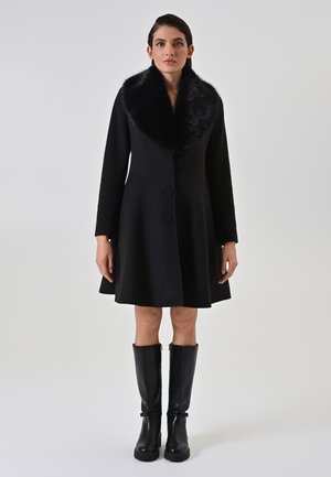 Schwarzer Mantel mit weitem Kunstfellkragen, tailliertem Oberteil, ausgestelltem Rock und drei großen Knöpfen. Das Model trägt außerdem kniehohe schwarze Stiefel.