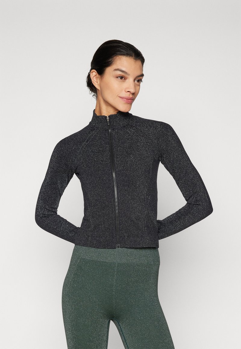 Sort zip-up sportsjakke med en glat, struktureret finish og en tætsiddende pasform. Båret over grønne leggings, med høj krave og lange ærmer.
