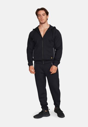Mężczyzna stojący, ubrany w czarną bluzę z zamkiem, dopasowane dresy i czarne buty sportowe na jednolitym tle.