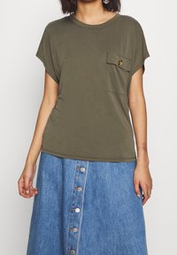 Olijfgroene T-shirt met korte mouwen, een borstzak en een knopdetail. Gecombineerd met een blauwe denimrok met knopen aan de voorkant.