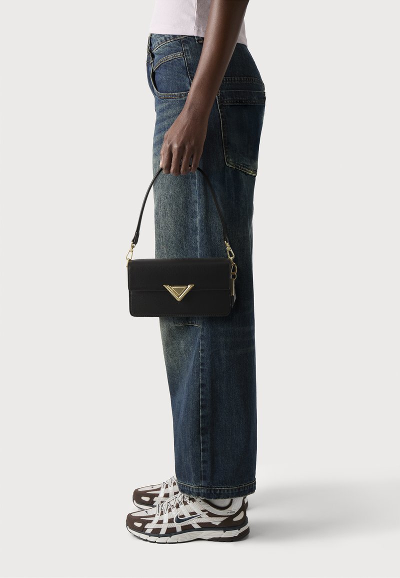 Black leather handbag with a triangular gold accent, rectangular shape, and short handle, being held next to blue denim jeans.