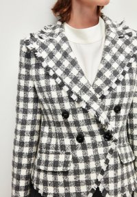 Blazer de cuadro de doble botonadura en blanco y negro, con bordes deshilachados, solapas de muesca y botones negros, que cuenta con dos bolsillos frontales.