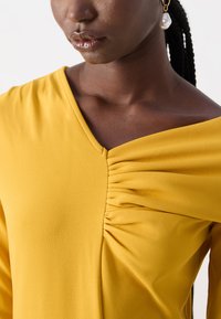 Keltainen paita, jossa on V-pääntie ja ryppydetaljointi vartalon kohdalla. Sujuva, kevyt kangas. Off-shoulder-leikkaus rentoa istuvuutta varten.