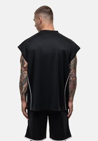 Zwart mouwloos sporttop met een losse fit, textuuroptiek en witte zijaccenten, gedragen met bijpassende shorts, waarbij tatoeages op de armen zichtbaar zijn.