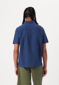 Short-sleeved shirt in dark blue linen, with a collar and yoke. Back features a straight hem and side slits. Worn with olive green pants.