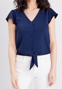 Navy blue lyhythihainen paita V-kaula-aukolla, edessä nappikiinnitys ja solmittu solmuhelma. Rakennekangas, jossa on hienovaraisia kuvioita.