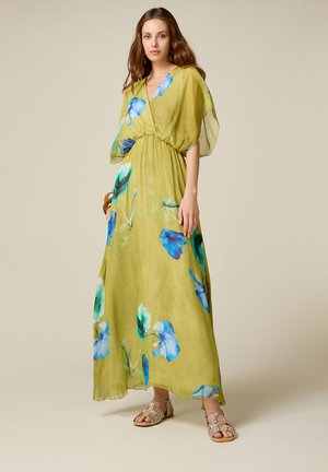 Kvinde iført en grøn, gulvlængde kjole med blå blomsterprint, stående mod en ensfarvet beige baggrund, iført sandaler og armbånd.