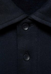 Námornícky polo tričko s texturovanou tkaninou, vyznačujúce sa špicatým golierom, dvoma čiernymi gombíkmi a detailným pohľadom na légu.