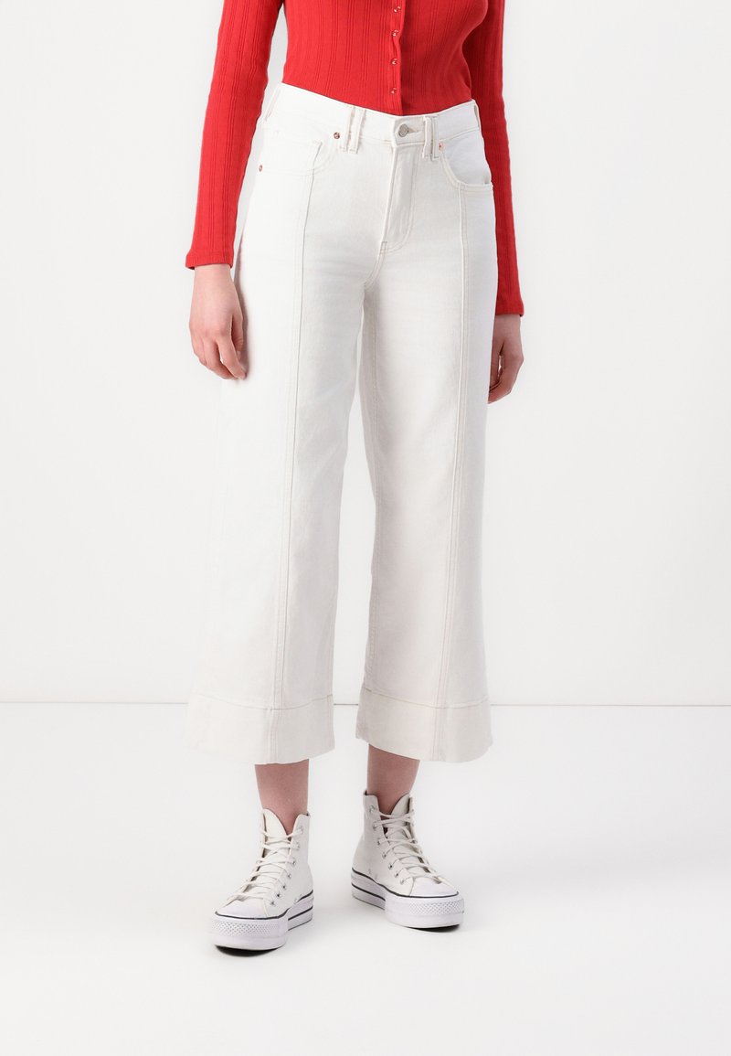 Vita vida jeans med hög midja, synliga sömmar och cropped längd, kombinerat med vita högskor och en röd långärmad tröja.