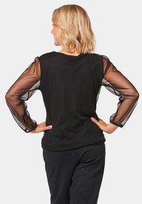 Schwarzes Oberteil mit transparenten langen Ärmeln, Rundhalsausschnitt und lockerer Passform. Die transparenten Ärmel haben ein Manschetten-Detail. Kombiniert mit dunkel gemusterten Hosen.