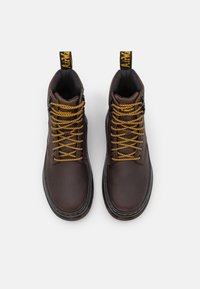 Botas de tobillo de cuero marrón con cordones amarillos y acentos negros. Presentan una punta redonda y una lengüeta con la marca en la parte superior. Superficie texturizada.