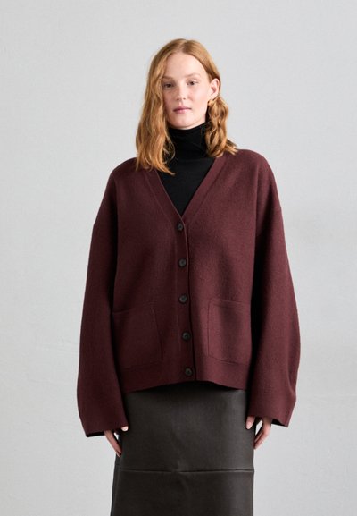 Burgunderfarbener Wollcardigan mit V-Ausschnitt, Frontknöpfen und zwei großen Taschen. Kombiniert mit einem schwarzen Rollkragenpullover und einem Lederrock.